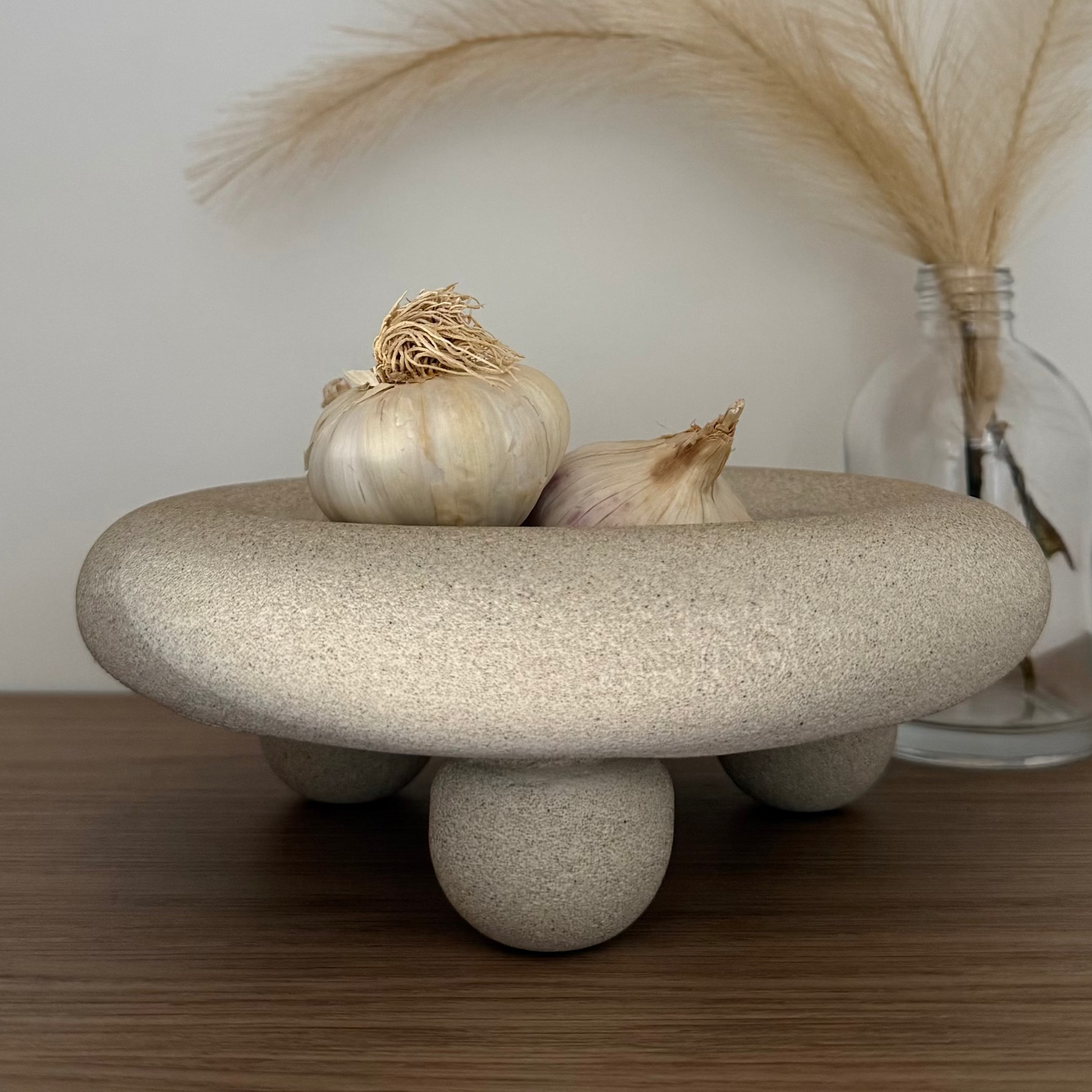 Handcrafted Halo Footed Bowl featuring a warm natural stone finish and rounded sculptural feet, styled with garlic on a tieber surface beside a glass bottle of dried pampas. 