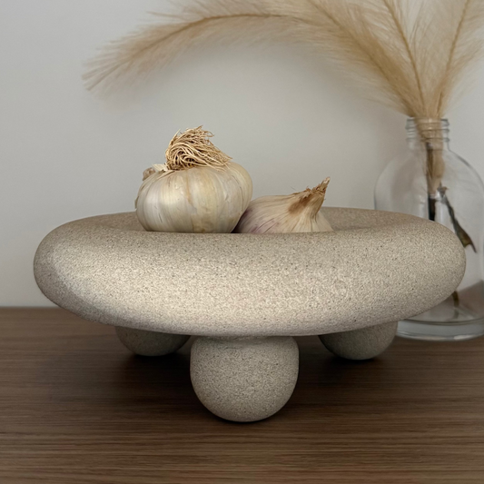 Handcrafted Halo Footed Bowl featuring a warm natural stone finish and rounded sculptural feet, styled with garlic on a tieber surface beside a glass bottle of dried pampas. 