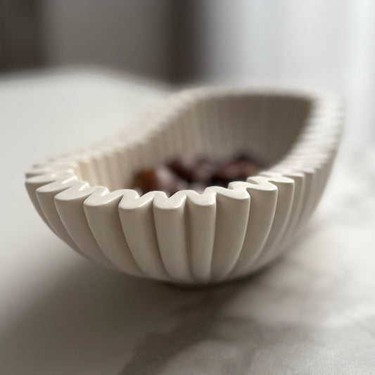 Handcrafted white ceramic bowl with a fluted, scalloped rim, shown on a light marble surface with blurred dark objects inside.